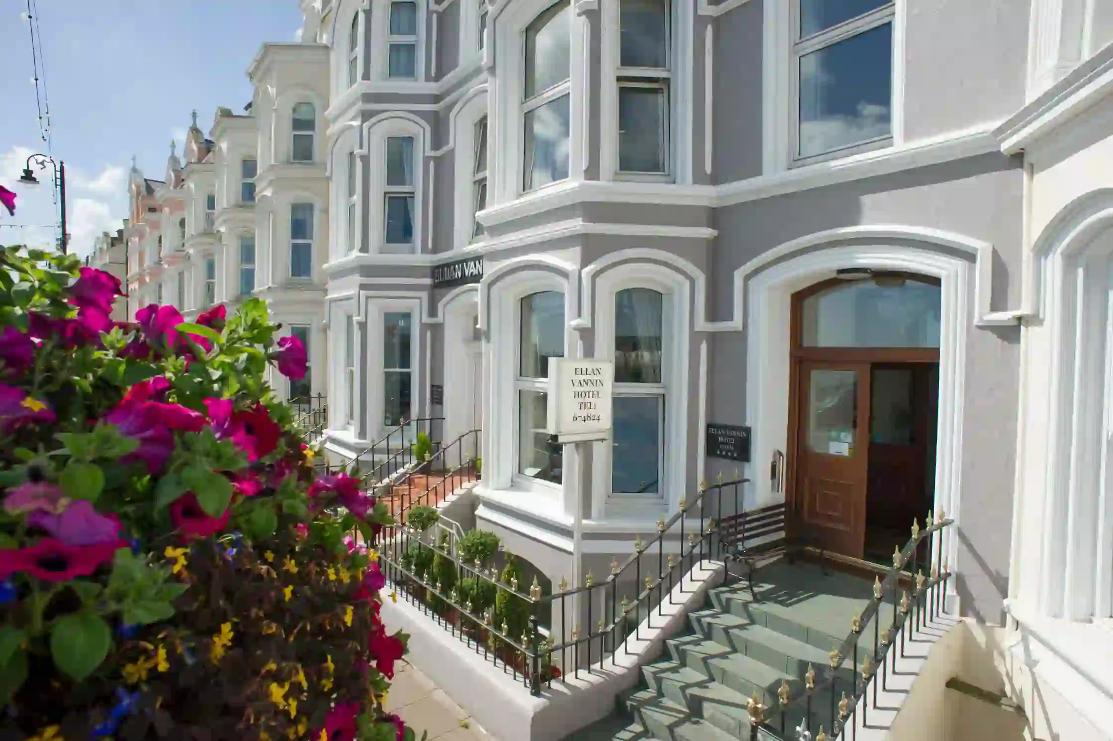Front entrance of the Ellan Vannin Hotel, a traditional building with bay windows, grey and white exterior, decorative railings, and vibrant purple flowers in the foreground
