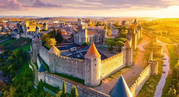 Bird's eye view of the Castle of Carcassonne at sunset, surrounded by trees and grass, and a pathway that loops around the castle. The castle has towers with red and blue turrets, and a distant view of a town can be seen behind it