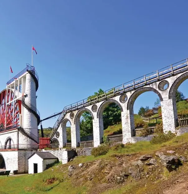 Low angle shot of the Laxey Wheel in the Isle of Man, which has a white base with a red wheel, on a grassy hill in front of a blue sky