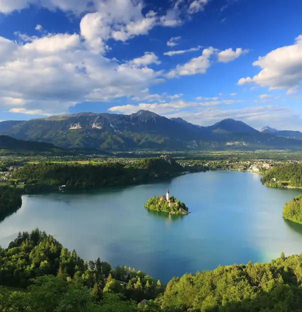 Wide angle shot of Lake Bled, showing its forested surrounding areas, and mountains in the distance. In the middle of the lake, there is a small island with trees and a towered building