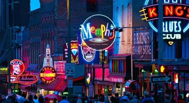 Vibrant night scene of Beale Street in Memphis, Tennessee, with colourful neon signs for bars, restaurants, and blues clubs, and a crowd of people walking along the street