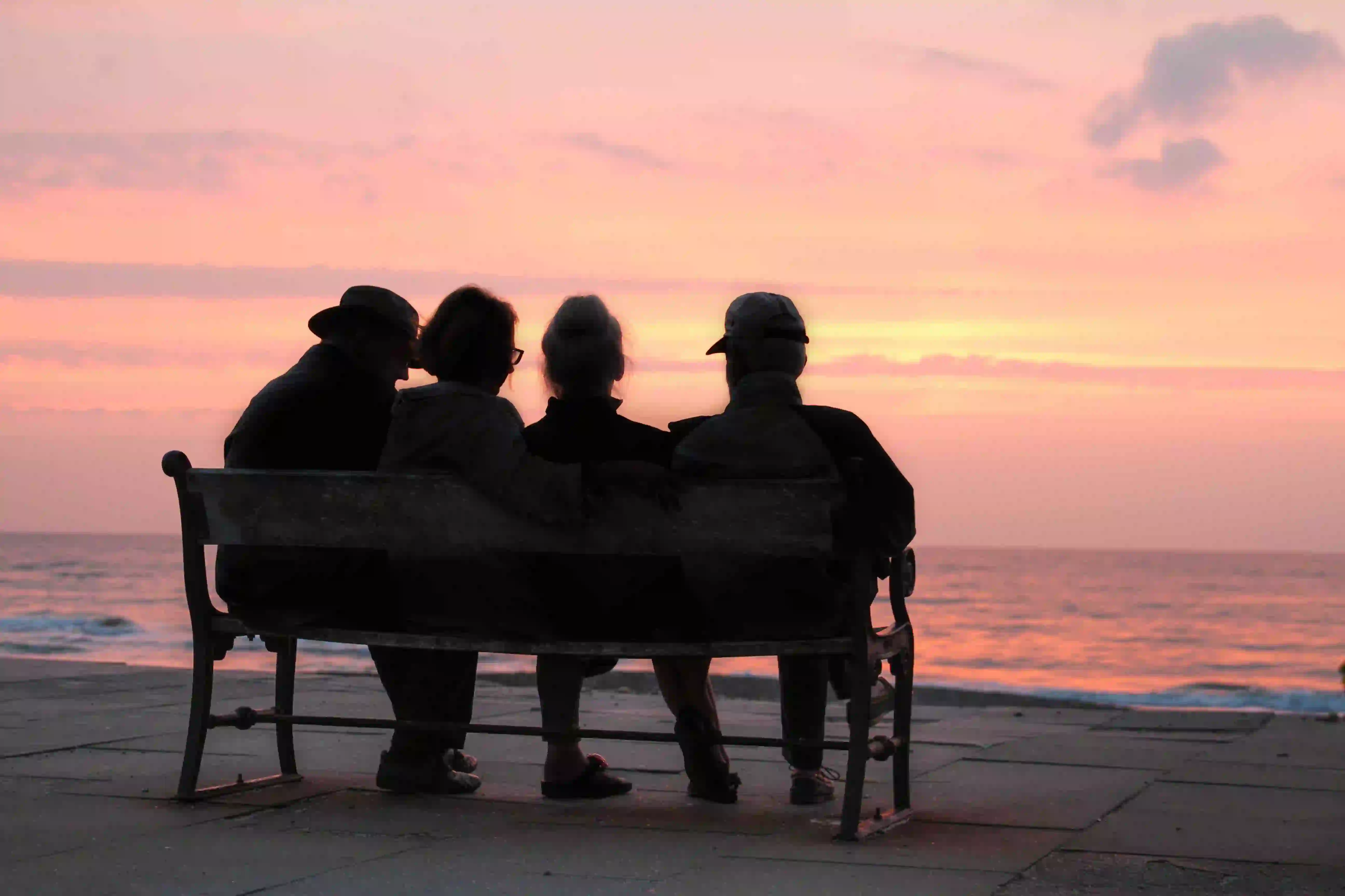 Over 70s friends sitting on a bench looking out at the sea at sunset