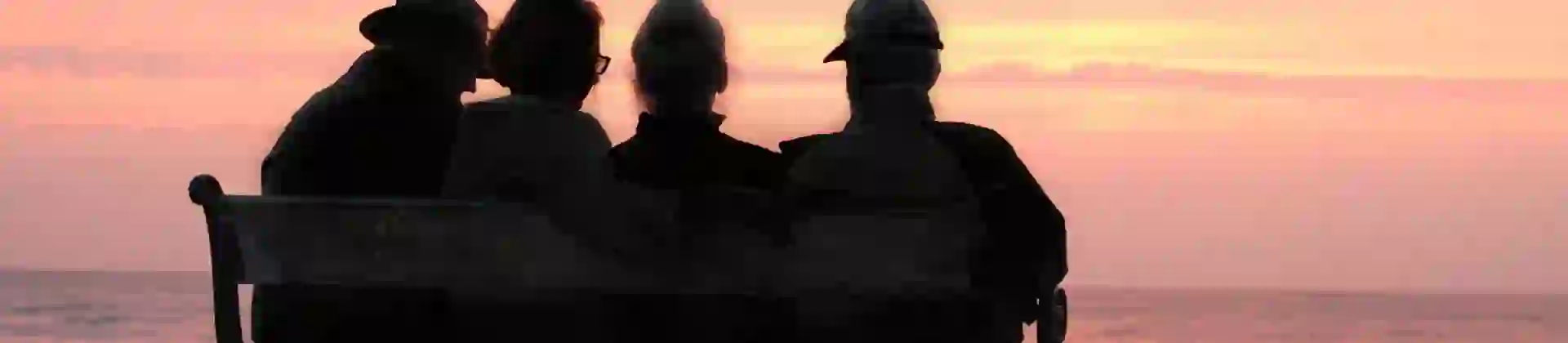 Over 70s friends sitting on a bench looking out at the sea at sunset