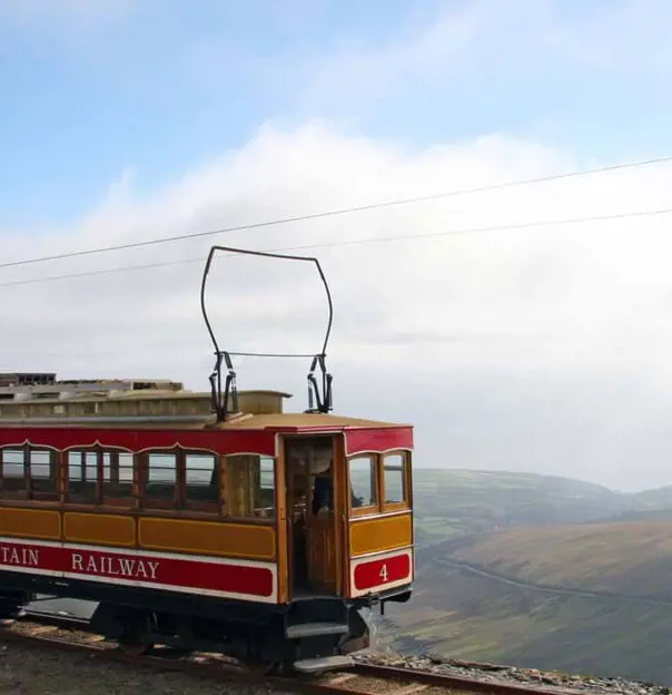 'Snaefell Mountain Railway' electric train travelling on the side of a mountain, with clouds and mountains behind it 