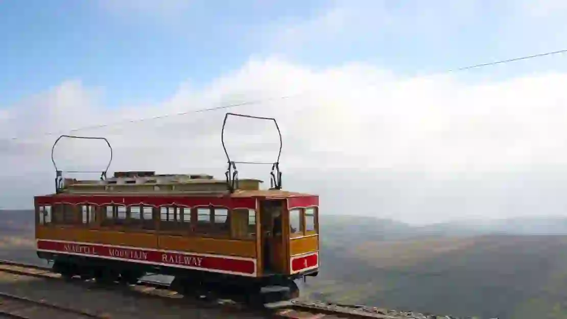 'Snaefell Mountain Railway' electric train travelling on the side of a mountain, with clouds and mountains behind it 