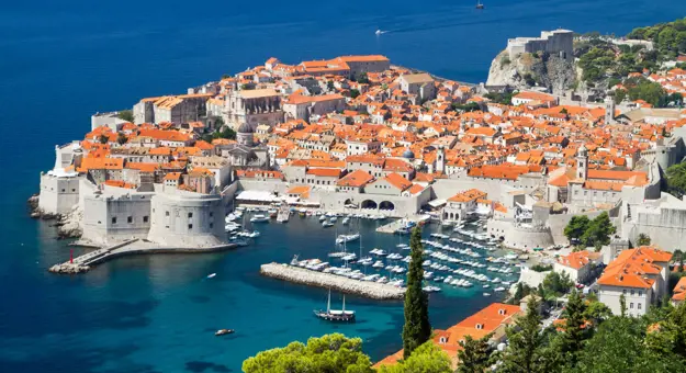 High angle view of Dubrovnik's coast and its harbour, with boats docked and lots of buildings with orange roofs behind. The navy blue sea to the left.