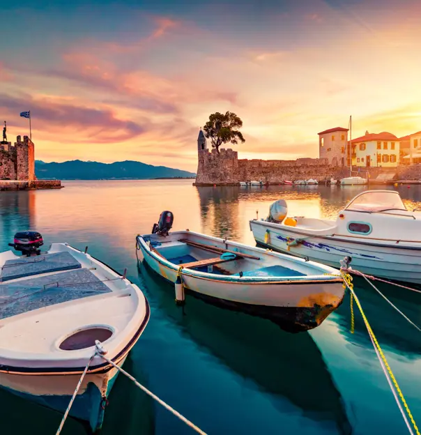 Small boats docked on the water, with a view of the ruins of a castle in the background, and mountains in the distance