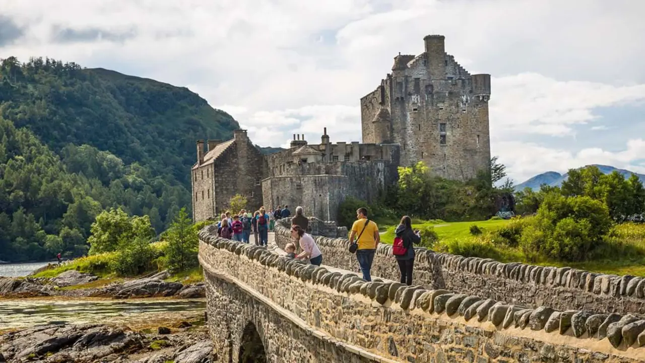 Eilean Donan Castle with people walking on the stone bridge walking towards it, and a mountain covered in trees behind it