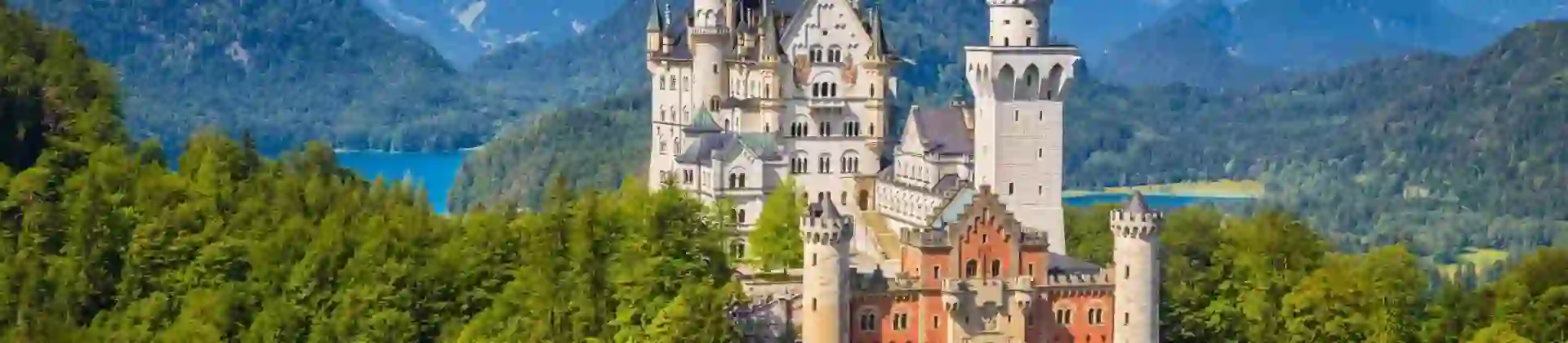 Neuschwanstein Castle in Bavaria, Germany, sitting atop a forested hill with mountain and a lake in the background