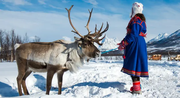 A lady dressed in traditional Norwegian clothes with a reindeer, standing in the snow