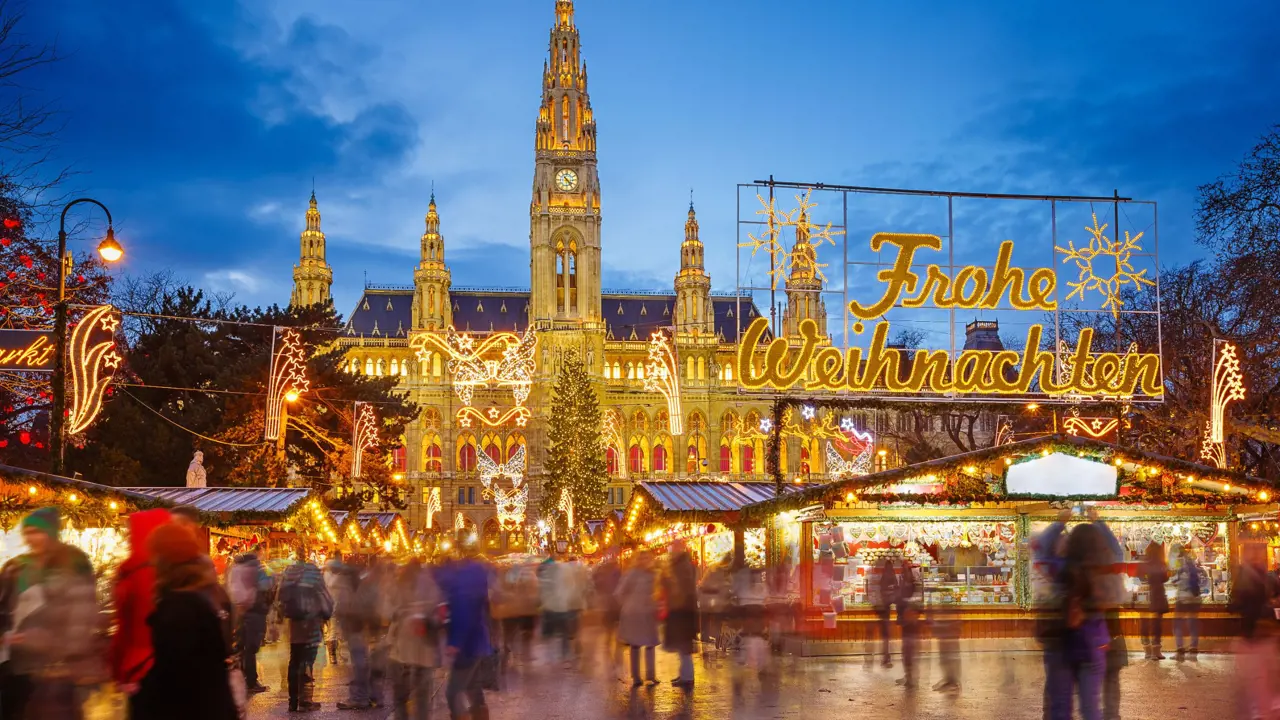 Shot of Vienna's gothic city hall which has one tall clock tower and four smaller towers, two on each side, all with pointy turrets. The building is a gold colour and is in front of a bright blue night sky. In front, is a Christmas Market, showing strips of cabin-like stalls and a variety of lit up Christmas decorations, one in the forefront reading 'Merry Christmas' in Austrian. People can be seen, although blurry as they are moving around. 