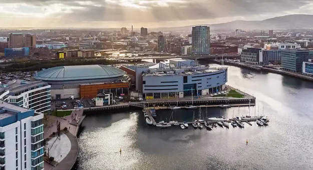 High angle view of Ulster Museum, a spherical, grey building on the edge of the River Lagan and next to a harbour with parked boats. The rest of the city of Belfast can be seen below a cloudy sky