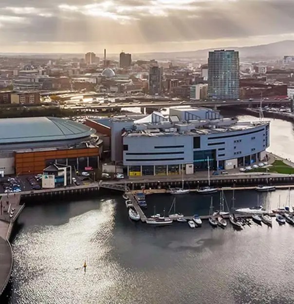 High angle view of Ulster Museum, a spherical, grey building on the edge of the River Lagan and next to a harbour with parked boats. The rest of the city of Belfast can be seen below a cloudy sky