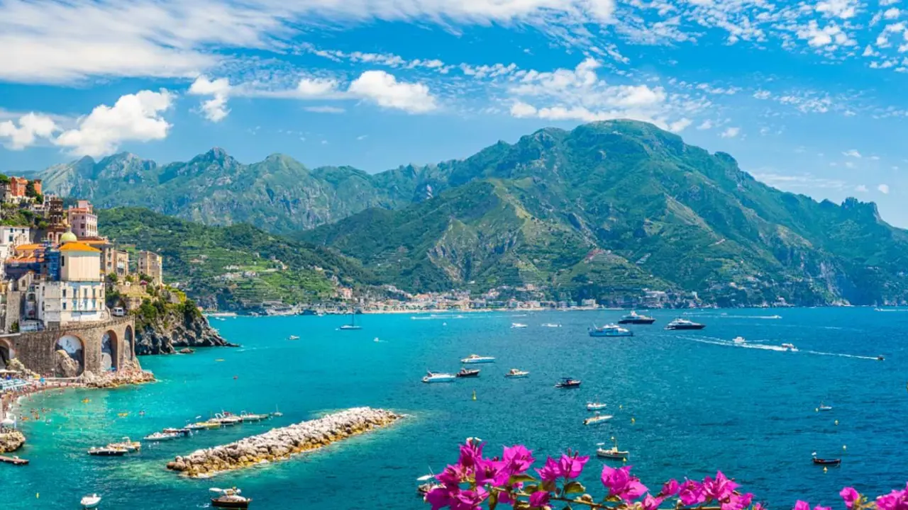 Scenic View Of Amalfi Coast Italy, showing the sea and boats, mountain in the distance, town on the edge of a cliff and pink flowers in the forefront