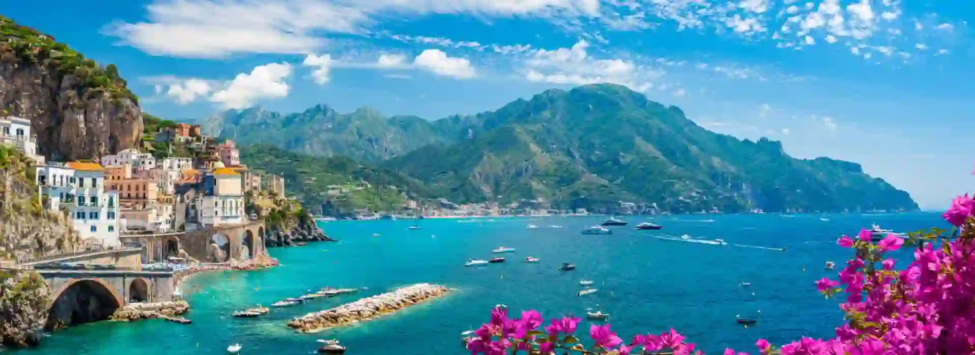 Scenic View Of Amalfi Coast Italy, showing the sea and boats, mountain in the distance, town on the edge of a cliff and pink flowers in the forefront