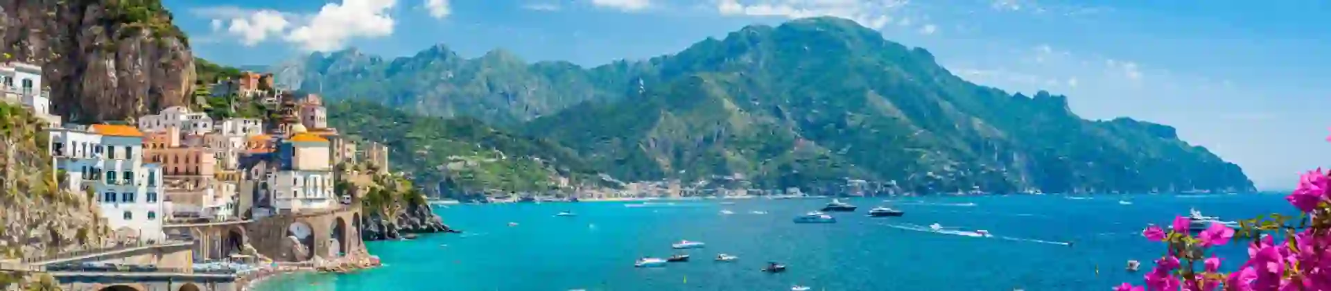 Scenic View Of Amalfi Coast Italy, showing the sea and boats, mountain in the distance, town on the edge of a cliff and pink flowers in the forefront