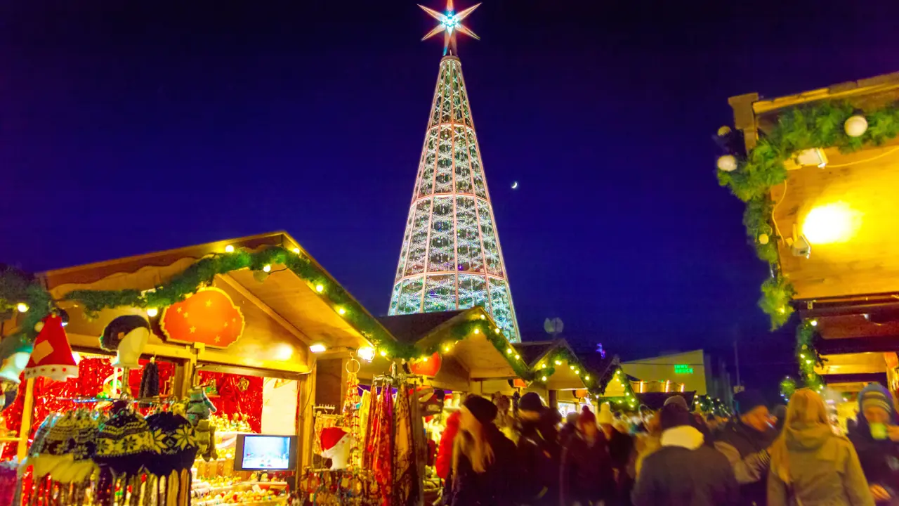 View of a Christmas market, with wooden, cabin-like stalls in the forefront, selling winter hats, dream catchers and scarves. The roofs are lined with green leaf-like garlands and fairy lights. Behind these is a Christmas tree metal structure, with a large, lit up star on top, in front of a navy blue night sky.