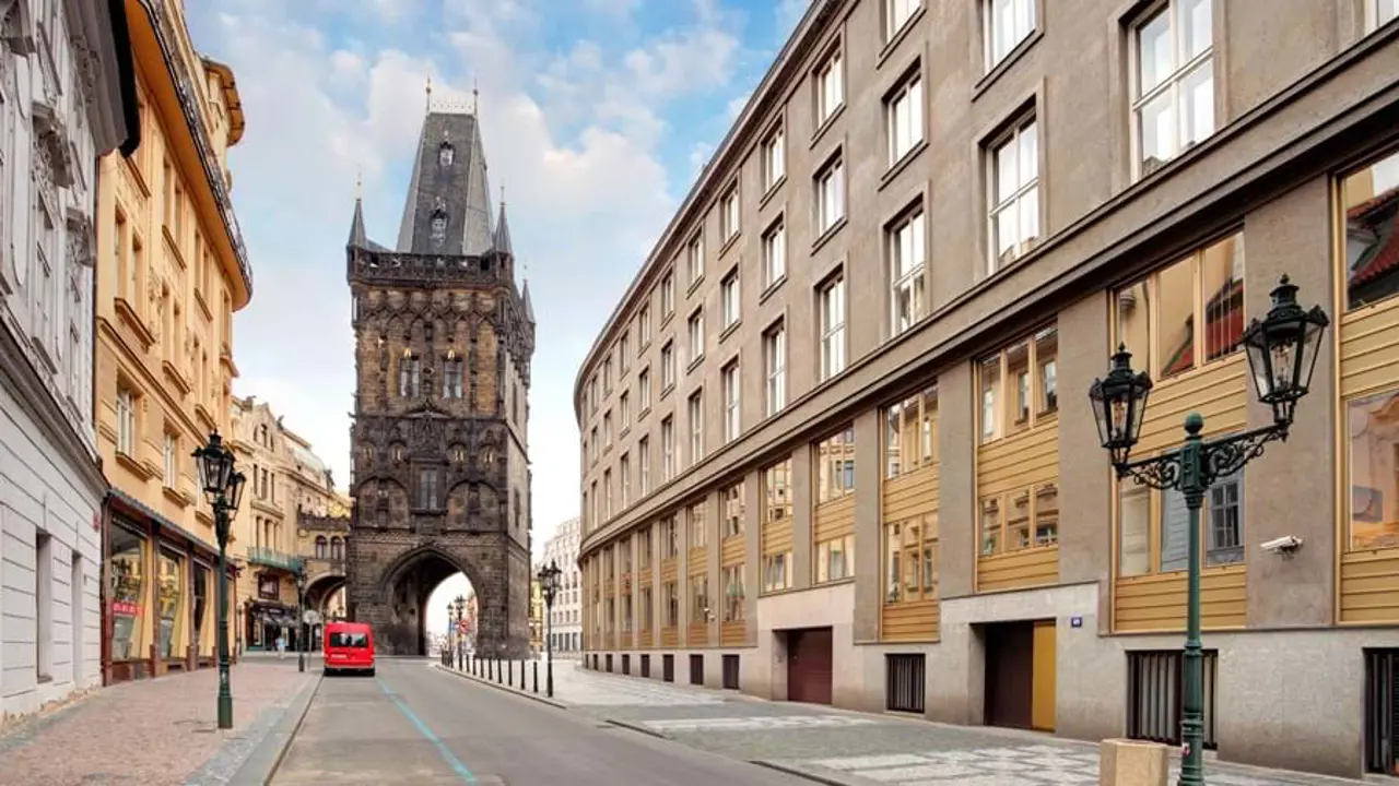 Shot of a street in Prague, buildings either side, and red car at the end, heading towards the archway of a dark-coloured tower with turrets. 