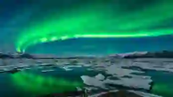 Green northern lights curving from the right back to the left. Below, a lake with ice blocks