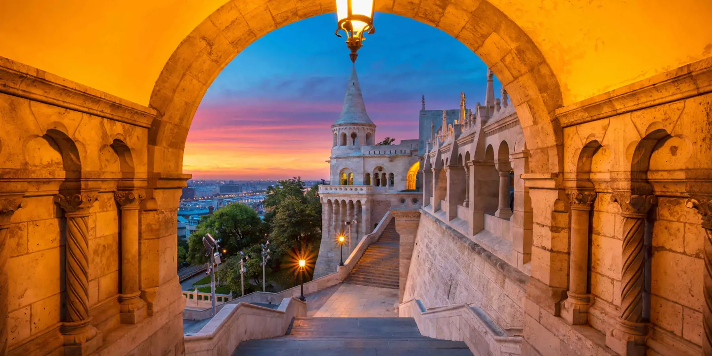 View of the side of a castle from an archway, showing a grey turret in front of blue, pink and orange sky