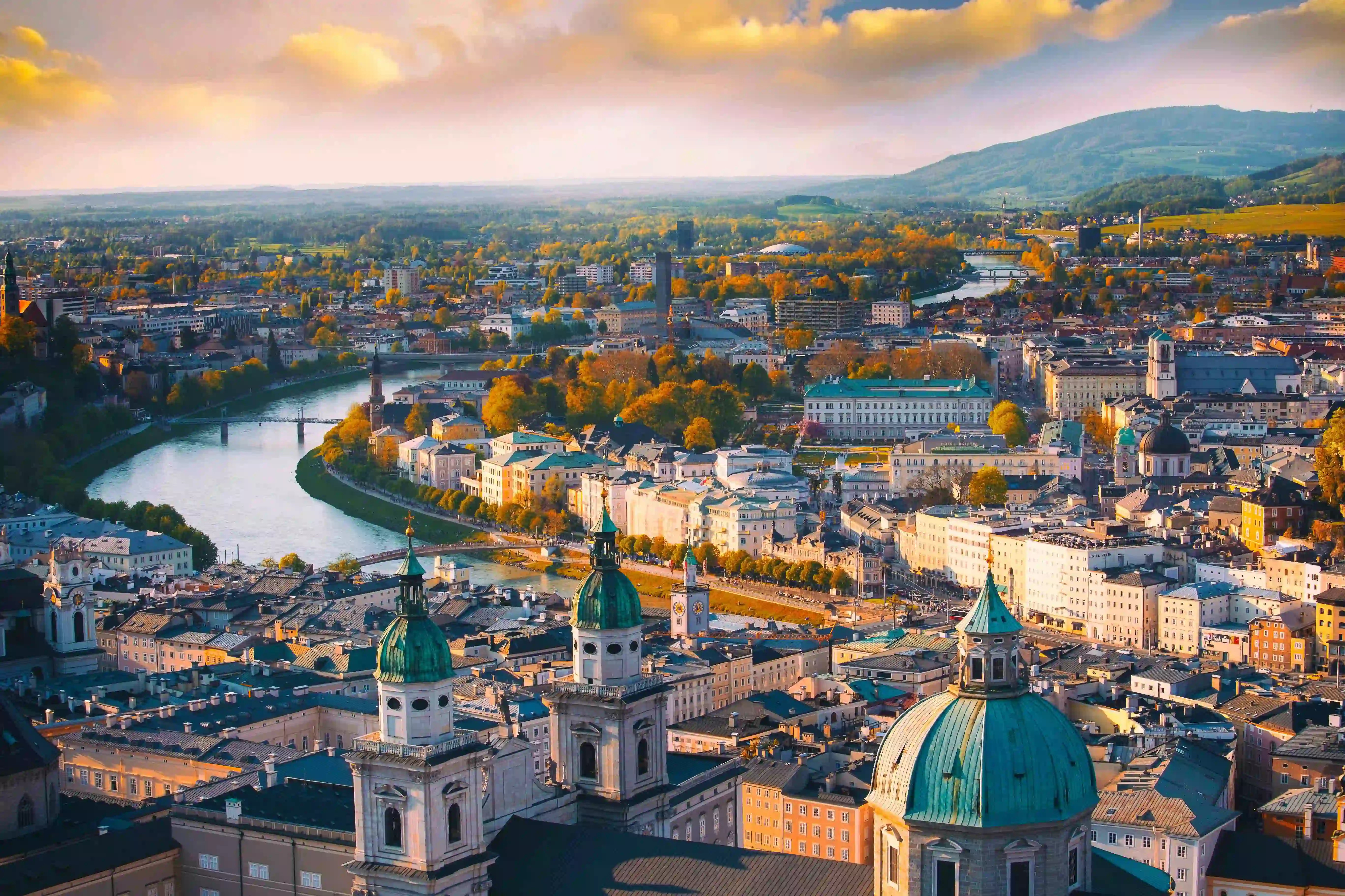 Bird's eye view of Salzburg, showing tall towers with turquoise turrets poking out of the top of rest of the buildings. In front of this you can see the river bending to the right and flowing into the distance. and the rest of the city's buildings and autumnal trees fill the majority of the image, going on into the distance, below a cloudy, slightly orange sky, and a mountain to the right.