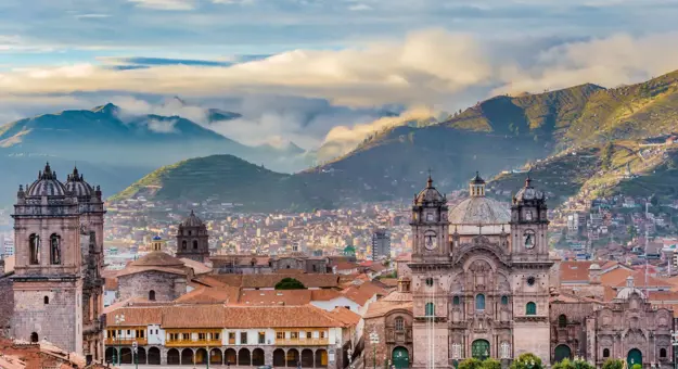 A panoramic view of Cusco, Peru, showcasing historic colonial buildings with terracotta roofs against a backdrop of green mountains and a cloudy sky.A panoramic view of Cusco, Peru, showcasing historic colonial buildings with terracotta roofs against a backdrop of green mountains and a cloudy sky