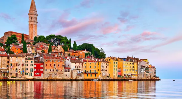 View of the town of Rovinj from the water, with colourful stone buildings on the waterfront, rows of these and trees going up the land behind and a tower with a statue on top. The sky is blue with pink clouds.