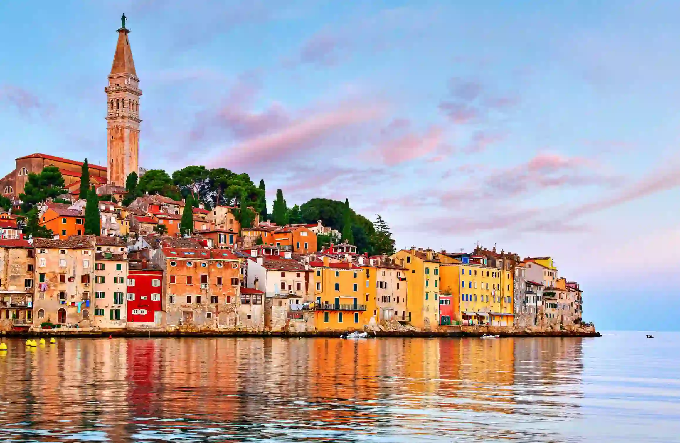 View of the town of Rovinj from the water, with colourful stone buildings on the waterfront, rows of these and trees going up the land behind and a tower with a statue on top. The sky is blue with pink clouds.