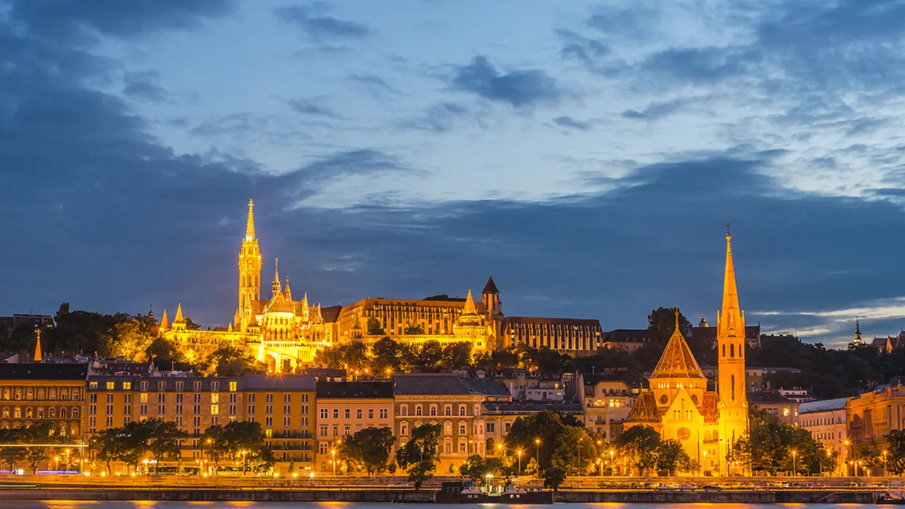 View of skyline of Buda, from Pest. With lit up buildings including two churches with spiked turrets.