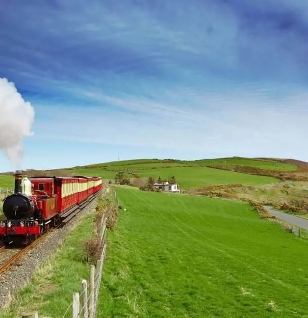 Red and black steam train heading towards the camera on a grassy hill on the coast of the Isle of Man, with a blue sky behind it