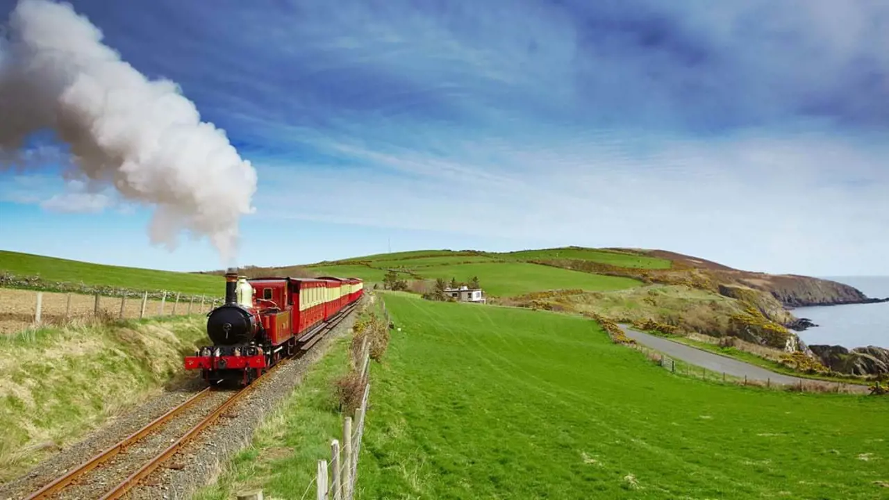 Red and black steam train heading towards the camera on a grassy hill on the coast of the Isle of Man, with a blue sky behind it