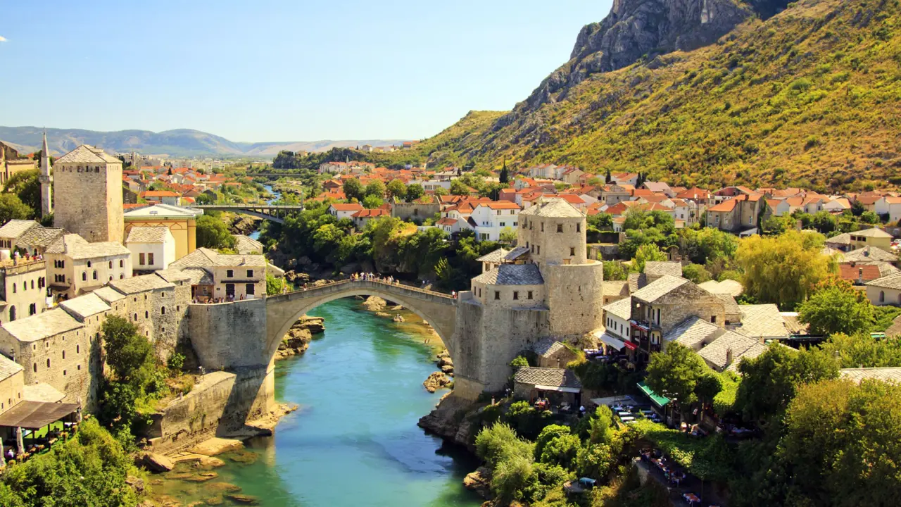 High angle view of Mostar Bridge, under is a turquoise river which flows into the distance. One the left are stone buildings and on the right some stone buildings and houses with orange roofs. Behind these, to the right of the image is the start of a mountain, covered in grass, and to the left is the blue sky.