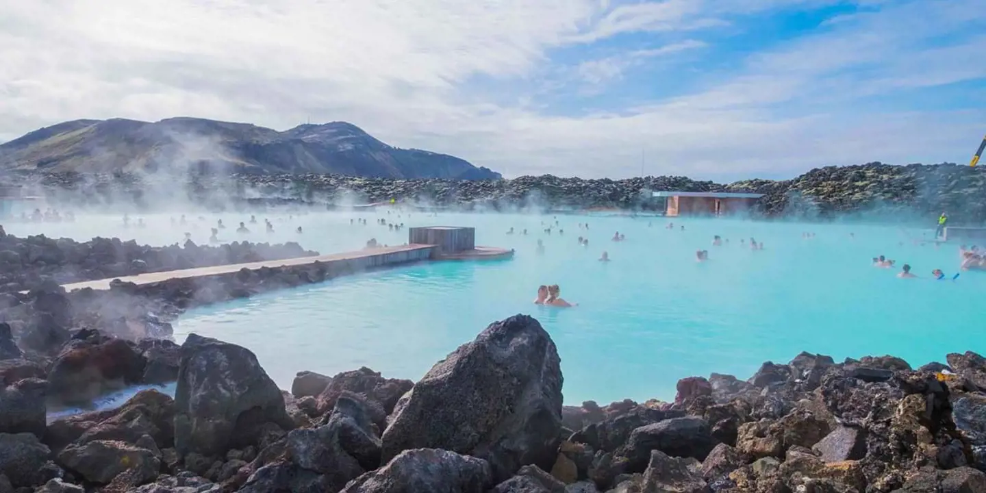 Geothermal lagoon in Iceland - bright blue water with steam coming off, surrounded by black rock. People swimming and mountains in the distance