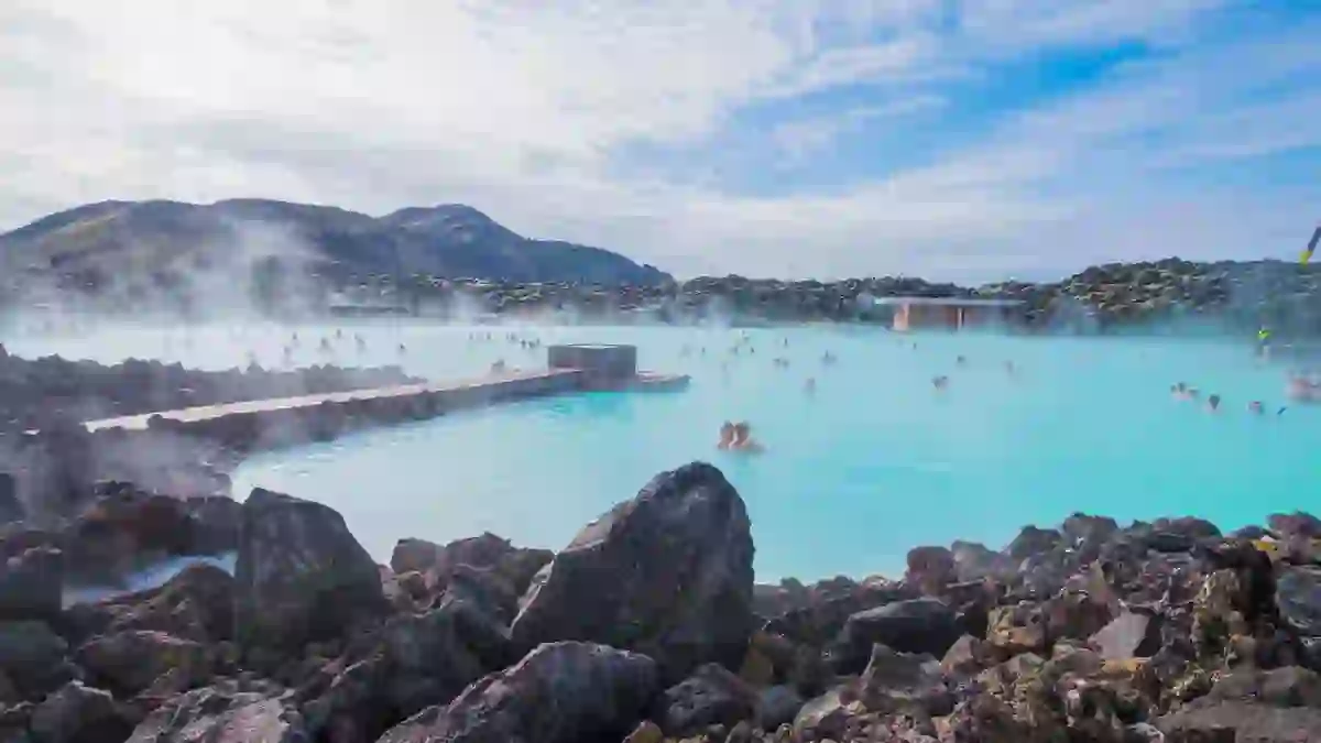 Geothermal lagoon in Iceland - bright blue water with steam coming off, surrounded by black rock. People swimming and mountains in the distance