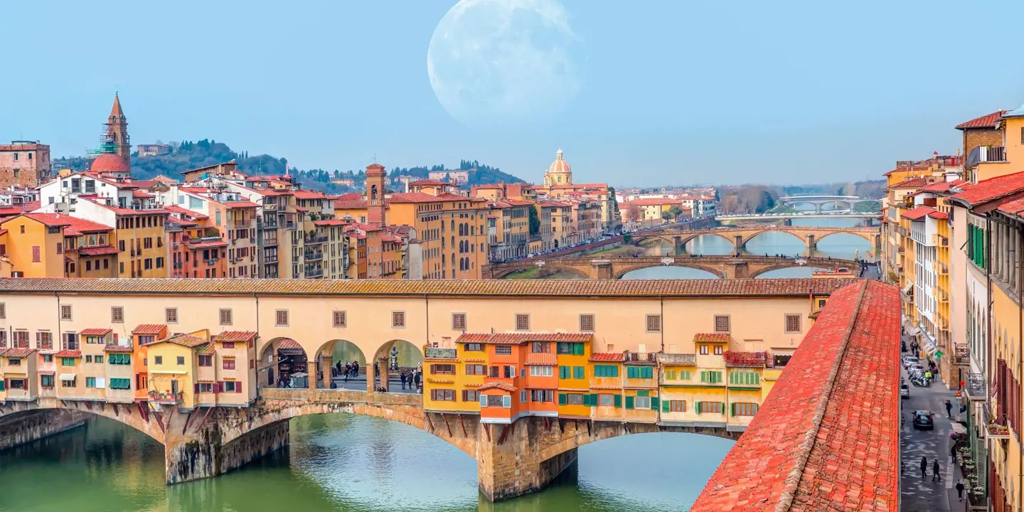 View of Ponte Vecchio Bridge showing buildings, the rest of The Arno River in the background, and a faint full moon in the sky