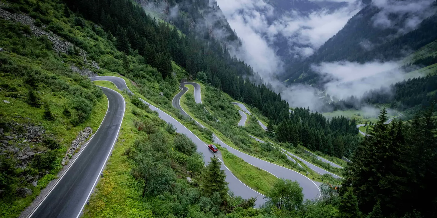 High angle shot of a road winding up a mountain in the Austrian Tyrol, with a red car on it. Fir trees cover the majority of the mountainsides, clouds further down the mountain and in the distance.