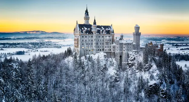 Wide angle shot Neuschwanstein Castle in the winter, showing its surrounding land covered in snow and a forest of fir trees in the forefront of the image. Behind the castle is a orange sky where the sun is setting