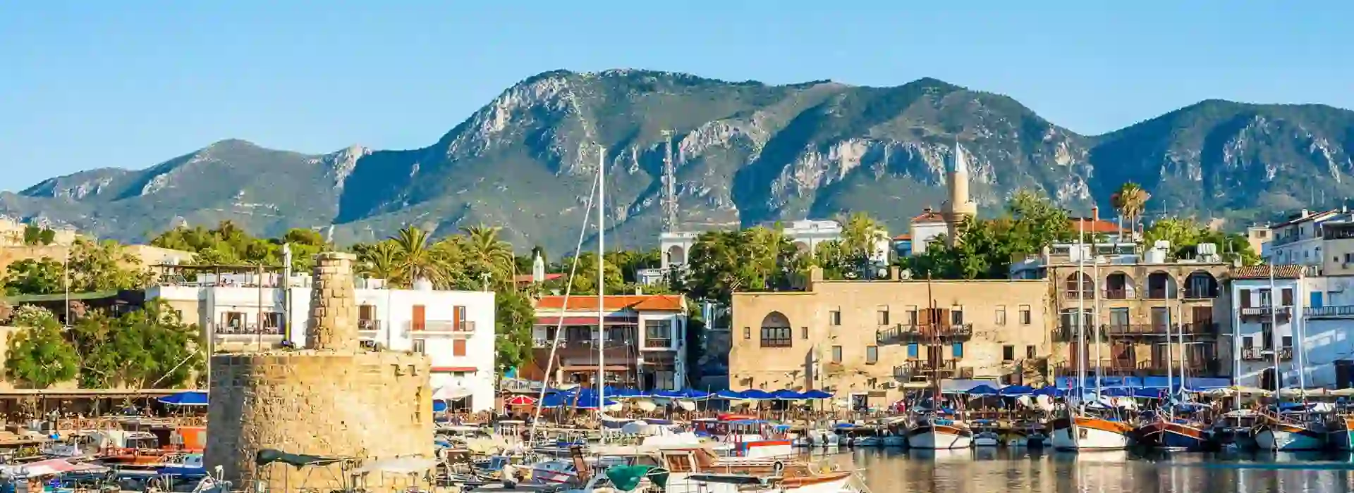 View of a harbour with boats docked, a ruin of a building on the left and buildings further back. Behind, a mountain and a blue sky.