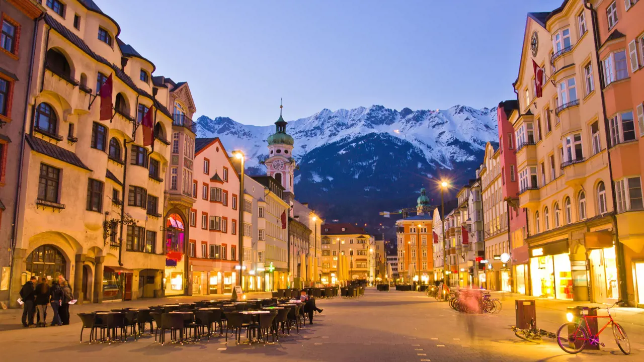 View of a wide road with shops and tall buildings either side. Tables and chairs are laid out in the road and bicycles are parked up on the right side. There is a clock tower which is taller than the rest of the buildings with a turquoise turret on the left, and snowy mountains can be seen at the end of the road, in front of light blue evening sky