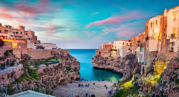 Image looking out to sea with some Polignano's buildings and a beach with people, at sunset