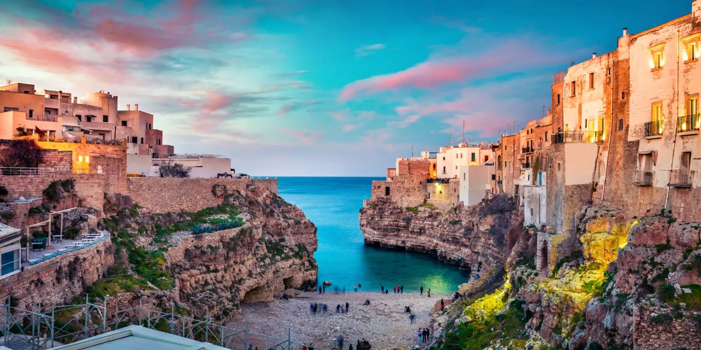 Image looking out to sea with some Polignano's buildings and a beach with people, at sunset