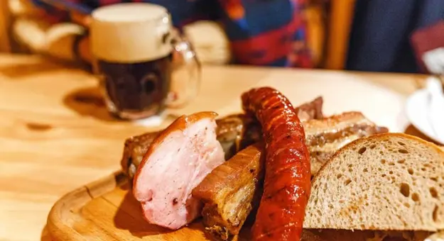 Close up of a plate of traditional sausage, pork meat and slices of bread.  Crafted Czech mug of beer in the background.