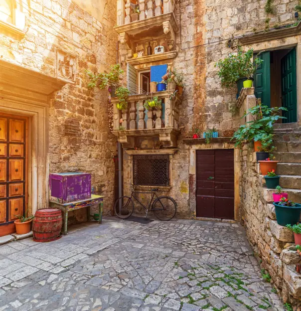 Shot of a concrete yard with steps on the right, with flower pots going up them, leading to a green door and a blue door. in the centre, a bicycle is parked up a stone wall, below two small Juliette balconies, then to the left there is a wide wooden door in a stone wall, and below it, empty flower pots, a table and a box.