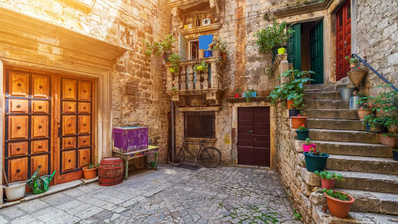 Shot of a concrete yard with steps on the right, with flower pots going up them, leading to a green door and a blue door. in the centre, a bicycle is parked up a stone wall, below two small Juliette balconies, then to the left there is a wide wooden door in a stone wall, and below it, empty flower pots, a table and a box.