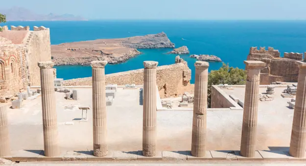 Stone pillars lined up in the forefront, with the ruins of Lindos city in front, and the view of the sea.