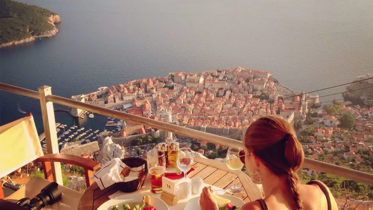 Back of a woman eating on a balcony overlooking Dubrovnik, the coastal town full of buildings with orange roofs on the seafront. On the table there is glasses of wine, a beer and bowls of salad