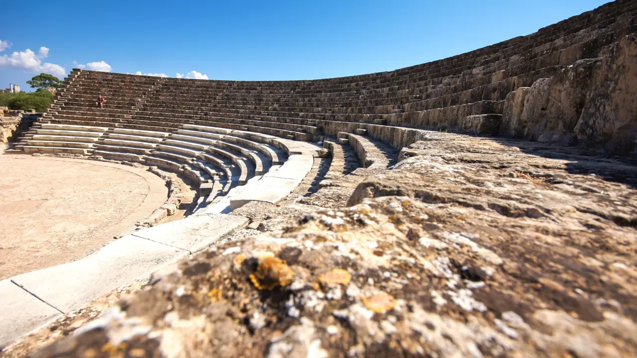 Low angle shot of the rows of seating in an ampitheatre, curving to the left, with a person sat on one of the rows on the far side. Above, a clear blue sky