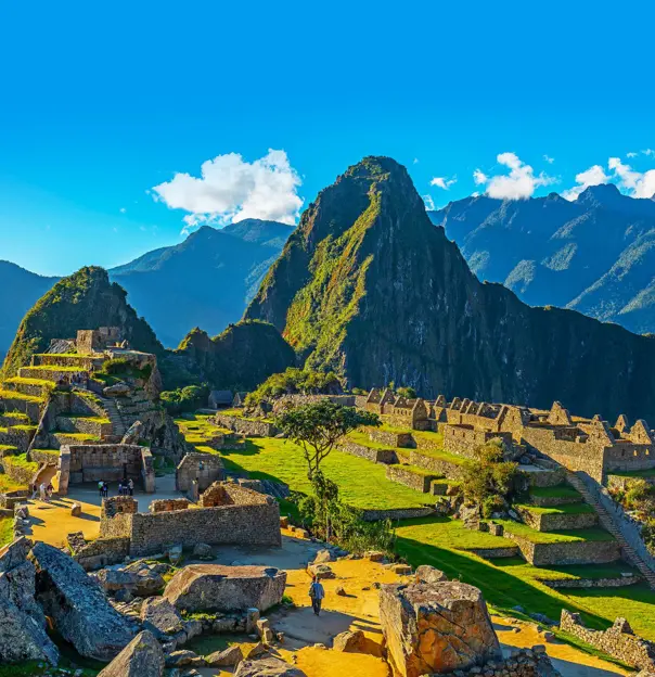 A panoramic view of Machu Picchu in Peru, showing ancient Incan ruins built into a green mountain ridge under a clear blue sky, with Huayna Picchu towering in the background
