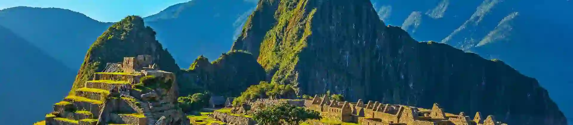 A panoramic view of Machu Picchu in Peru, showing ancient Incan ruins built into a green mountain ridge under a clear blue sky, with Huayna Picchu towering in the background