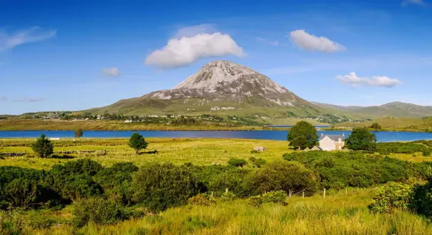 View of a mountain with fields in the forefront, and a river running in between. Blue sky with some clouds